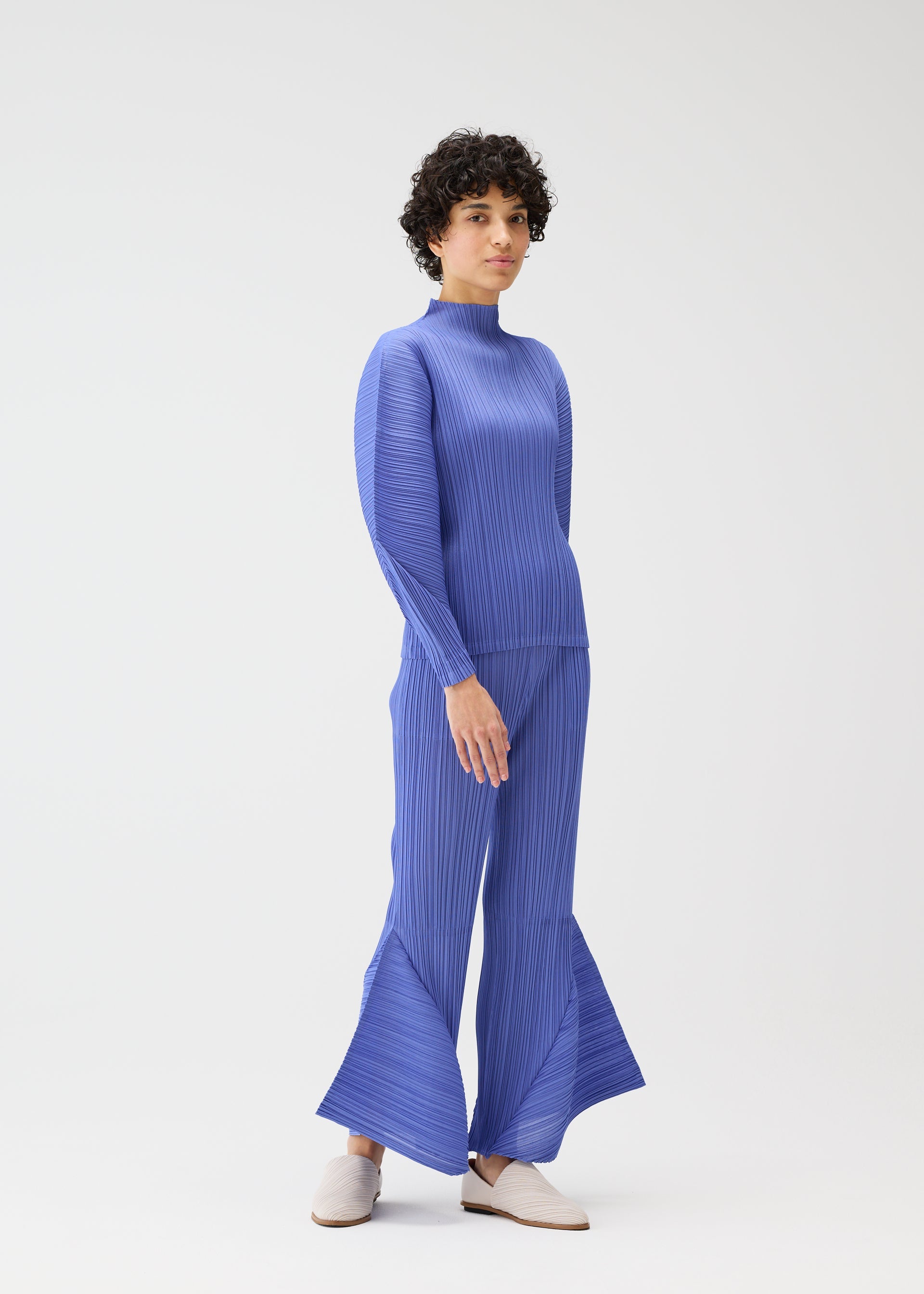 Zoomed view A person with short curly hair models the SKYLINE set by PLEATS PLEASE ISSEY MIYAKE: a textured blue long-sleeve top and flared city-inspired pants, styled with light slip-on shoes in a minimalist white studio.
