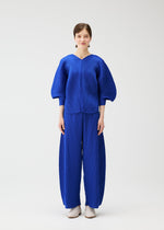 A woman stands facing forward against a white background, wearing the COAST two-piece wide pants and pleated sleeve set by PLEATS PLEASE ISSEY MIYAKE. Her short brown hair and white shoes complete the look.