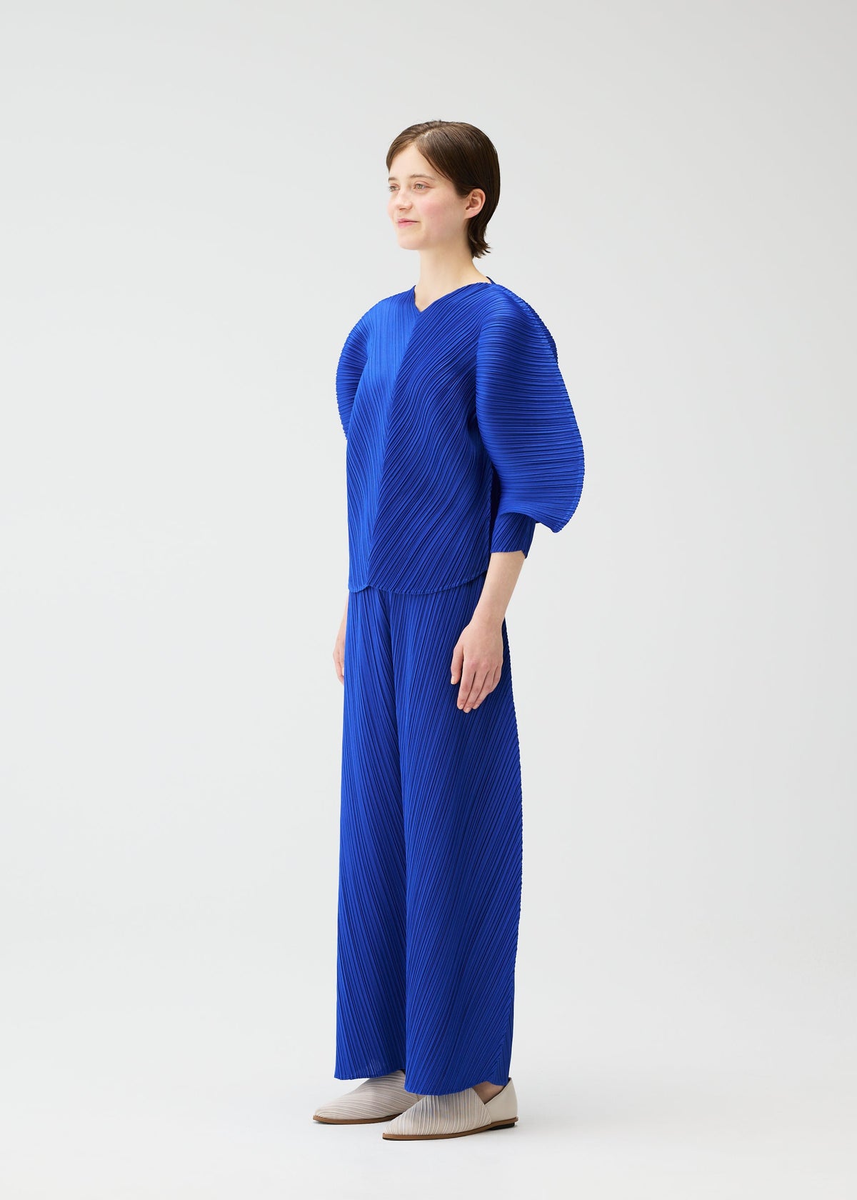 A person stands in a studio wearing the COAST set by PLEATS PLEASE ISSEY MIYAKE: a bright blue pleated top with rounded sleeves and matching diagonal-pleated wide pants, paired with neutral shoes against a white background. view 7 of 9