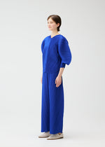 A person stands in a studio wearing the COAST set by PLEATS PLEASE ISSEY MIYAKE: a bright blue pleated top with rounded sleeves and matching diagonal-pleated wide pants, paired with neutral shoes against a white background.