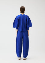 A person faces away, wearing the COAST set by PLEATS PLEASE ISSEY MIYAKE in cobalt blue. Diagonal pleats add texture to the voluminous sleeves and wide pants, set against a plain light background and paired with white shoes.