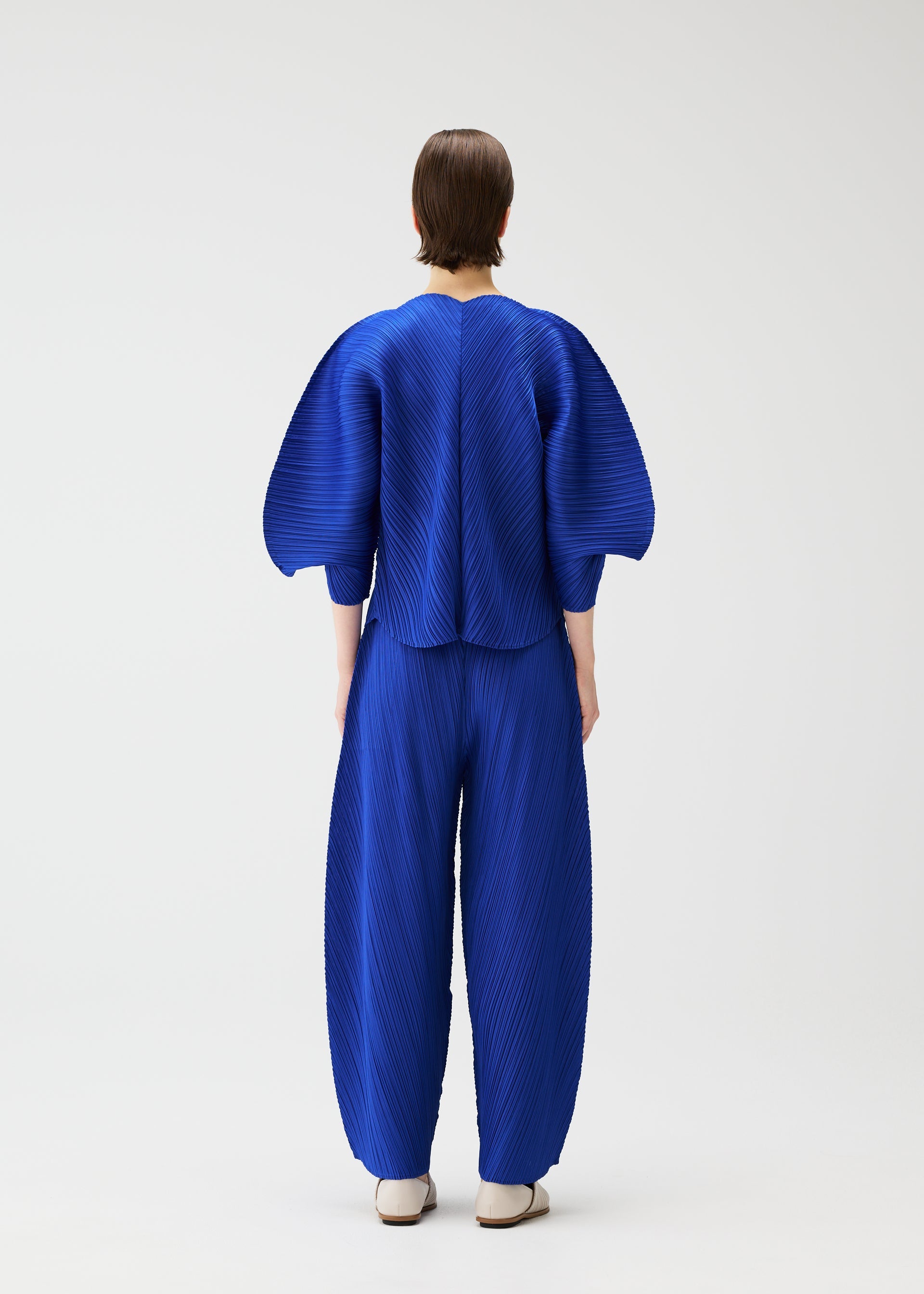 Zoomed view A person faces away, wearing the COAST set by PLEATS PLEASE ISSEY MIYAKE in cobalt blue. Diagonal pleats add texture to the voluminous sleeves and wide pants, set against a plain light background and paired with white shoes.