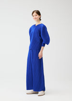 A person poses confidently against a plain white background, wearing the COAST vibrant blue outfit by PLEATS PLEASE ISSEY MIYAKE, with diagonal pleats, voluminous sleeves, wide pants, and white shoes. They smile slightly at the camera.