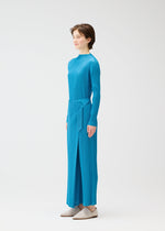 A person with short brown hair stands in profile against a white background, wearing the FACADE pleated top and matching wrap-detail pants by PLEATS PLEASE ISSEY MIYAKE, paired with light-colored shoes for a modern look.