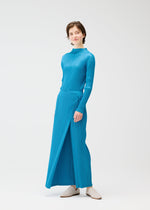 A model wears the FACADE dress by PLEATS PLEASE ISSEY MIYAKE—a bright blue, long-sleeved, pleated piece with high neckline and front slit—styled with white slip-on shoes, unconventional pants, and short brown hair against a plain white background.