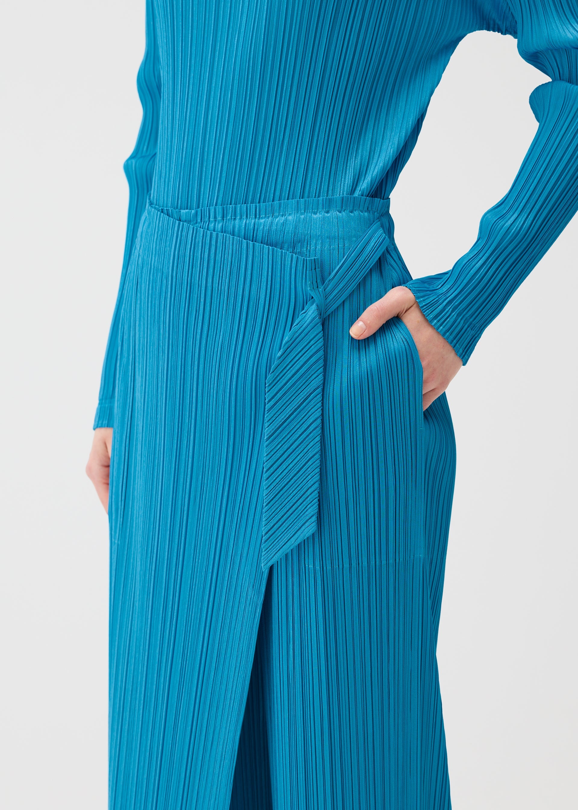 Zoomed view A person wears the FACADE set by PLEATS PLEASE ISSEY MIYAKE: a bright blue, pleated long-sleeve top and matching pants with a wrap detail, standing with one hand in their pocket against a plain background.