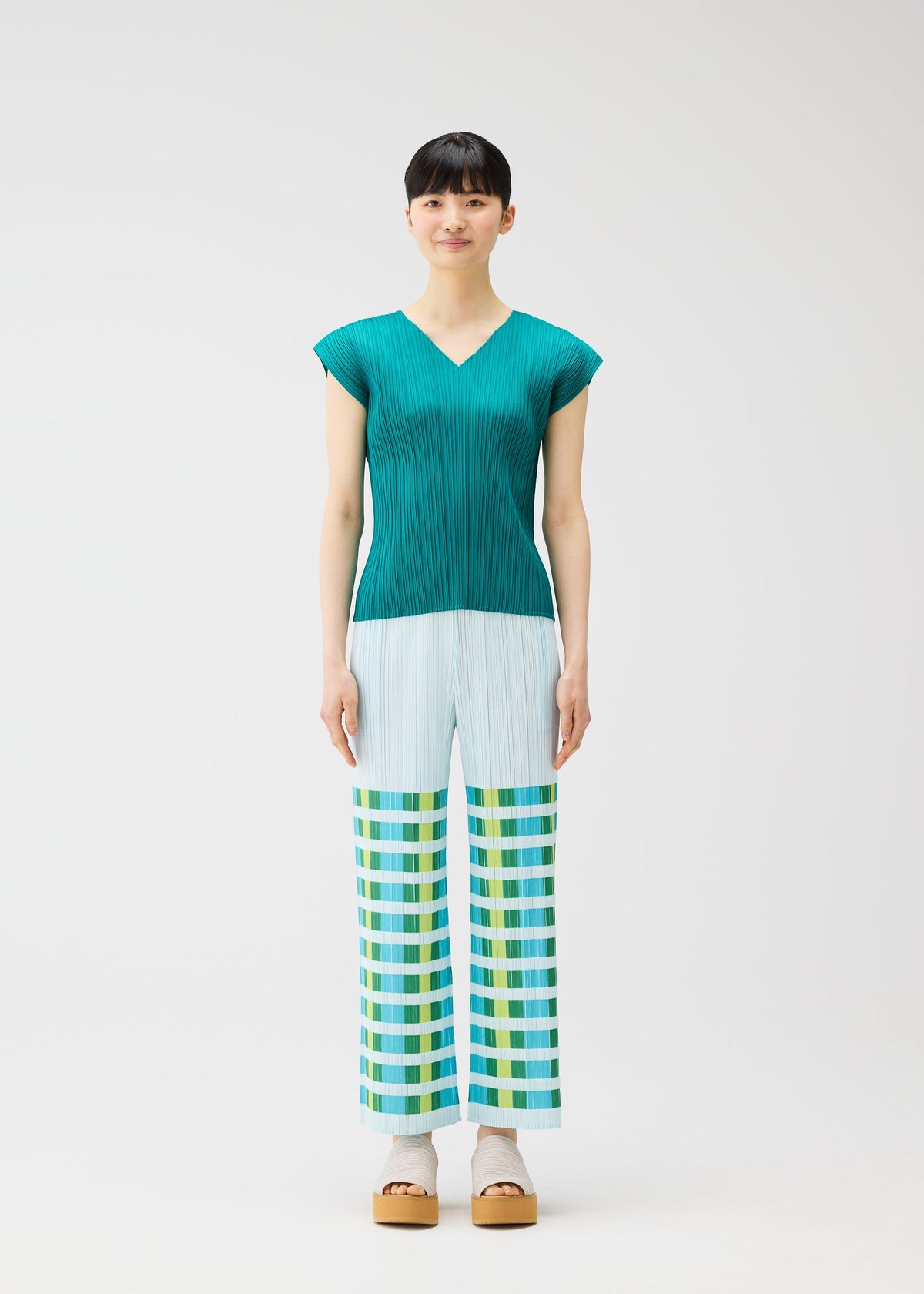 A woman faces forward in the BUMPY BASKET teal pleated short-sleeve top and light blue plaid straight pants with green, yellow, and blue geometric pattern by PLEATS PLEASE ISSEY MIYAKE. She wears beige platform sandals against a plain white background. view 5 of 8