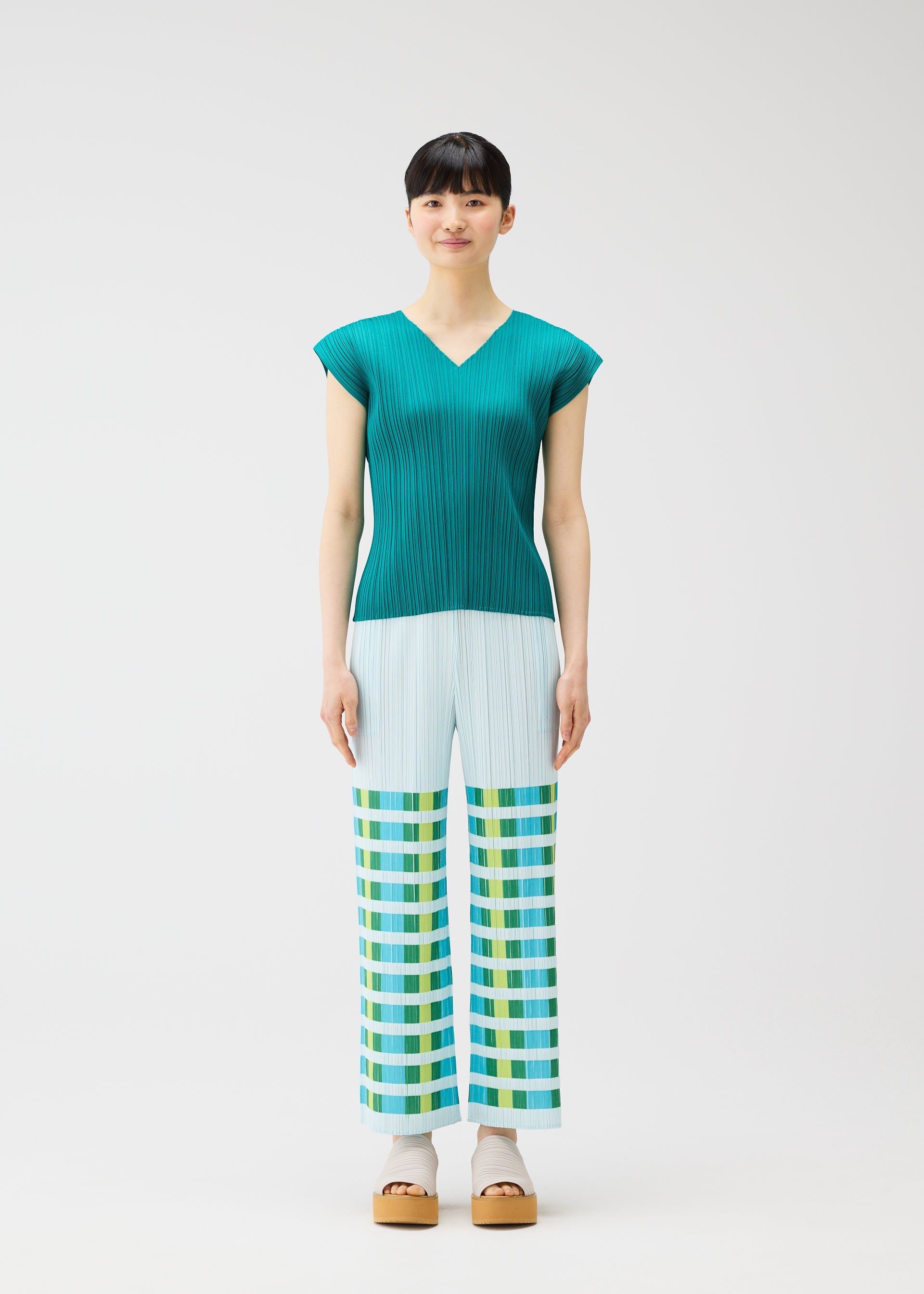 Zoomed view A woman faces forward in the BUMPY BASKET teal pleated short-sleeve top and light blue plaid straight pants with green, yellow, and blue geometric pattern by PLEATS PLEASE ISSEY MIYAKE. She wears beige platform sandals against a plain white background.