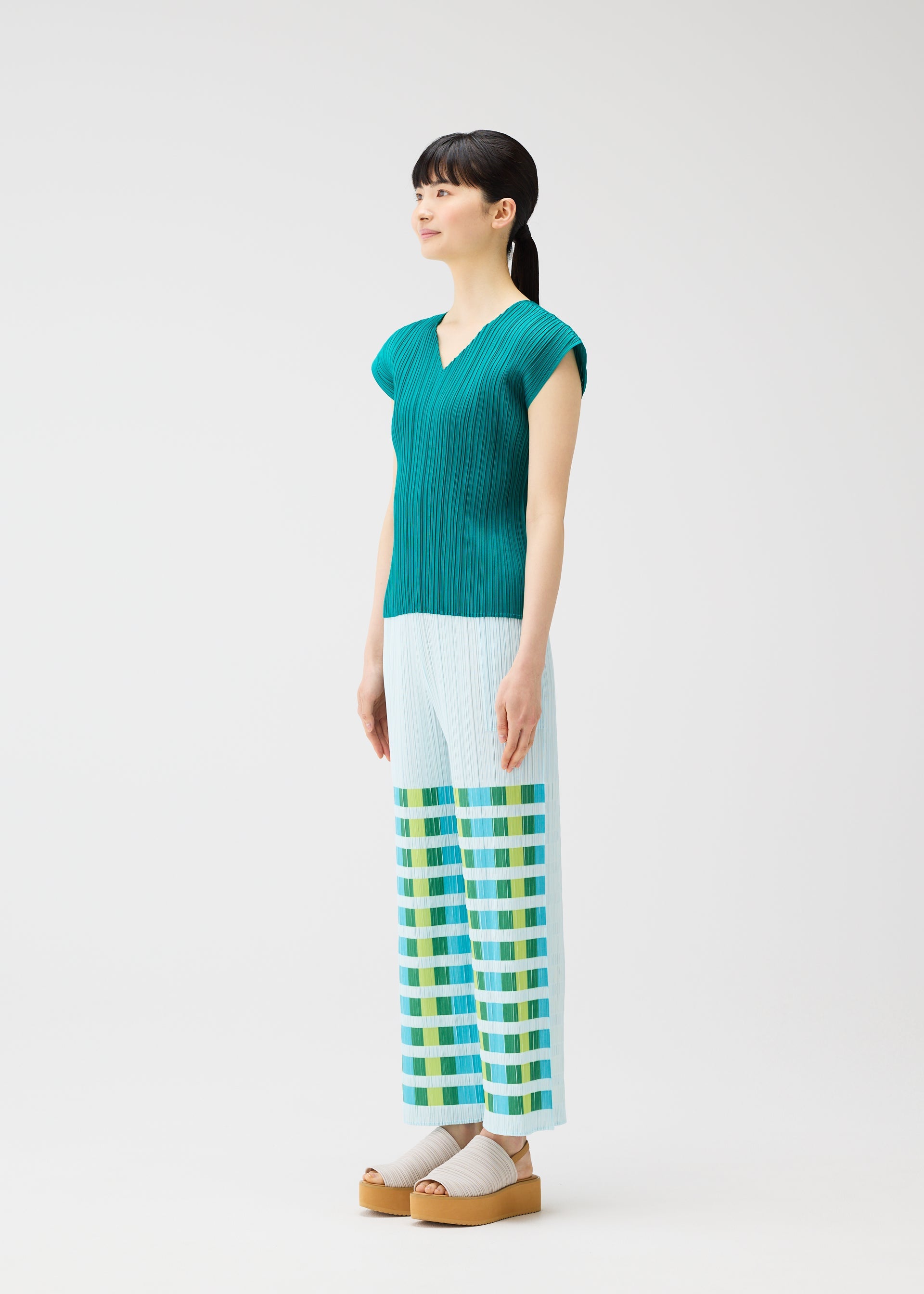 Zoomed view A woman with dark hair in a ponytail wears the BUMPY BASKET teal pleated blouse and plaid straight pants by PLEATS PLEASE ISSEY MIYAKE, featuring green and yellow checks near the hems, paired with beige platform sandals.