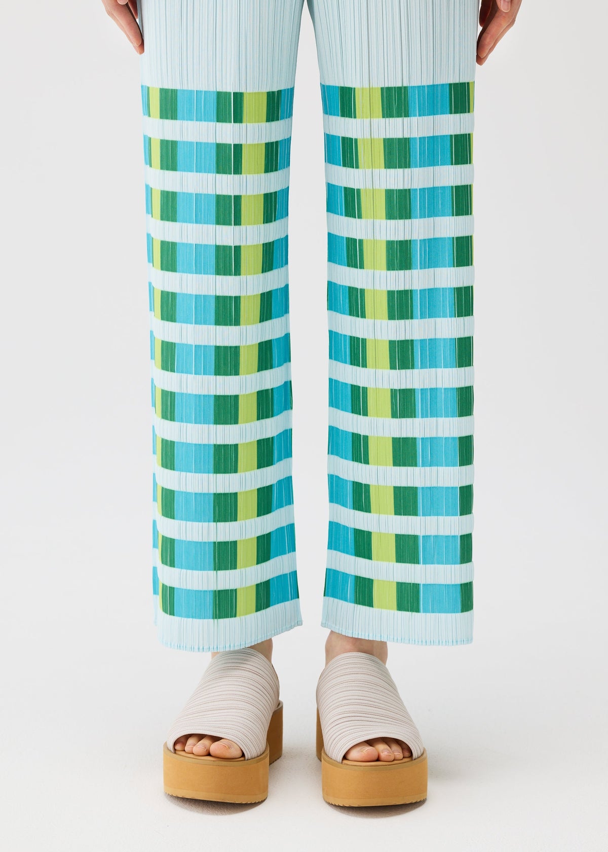 A person wears the PLEATS PLEASE ISSEY MIYAKE BUMPY BASKET plaid straight pants with green and turquoise stripes, styled with beige platform sandals with thick straps on a white background. view 8 of 8