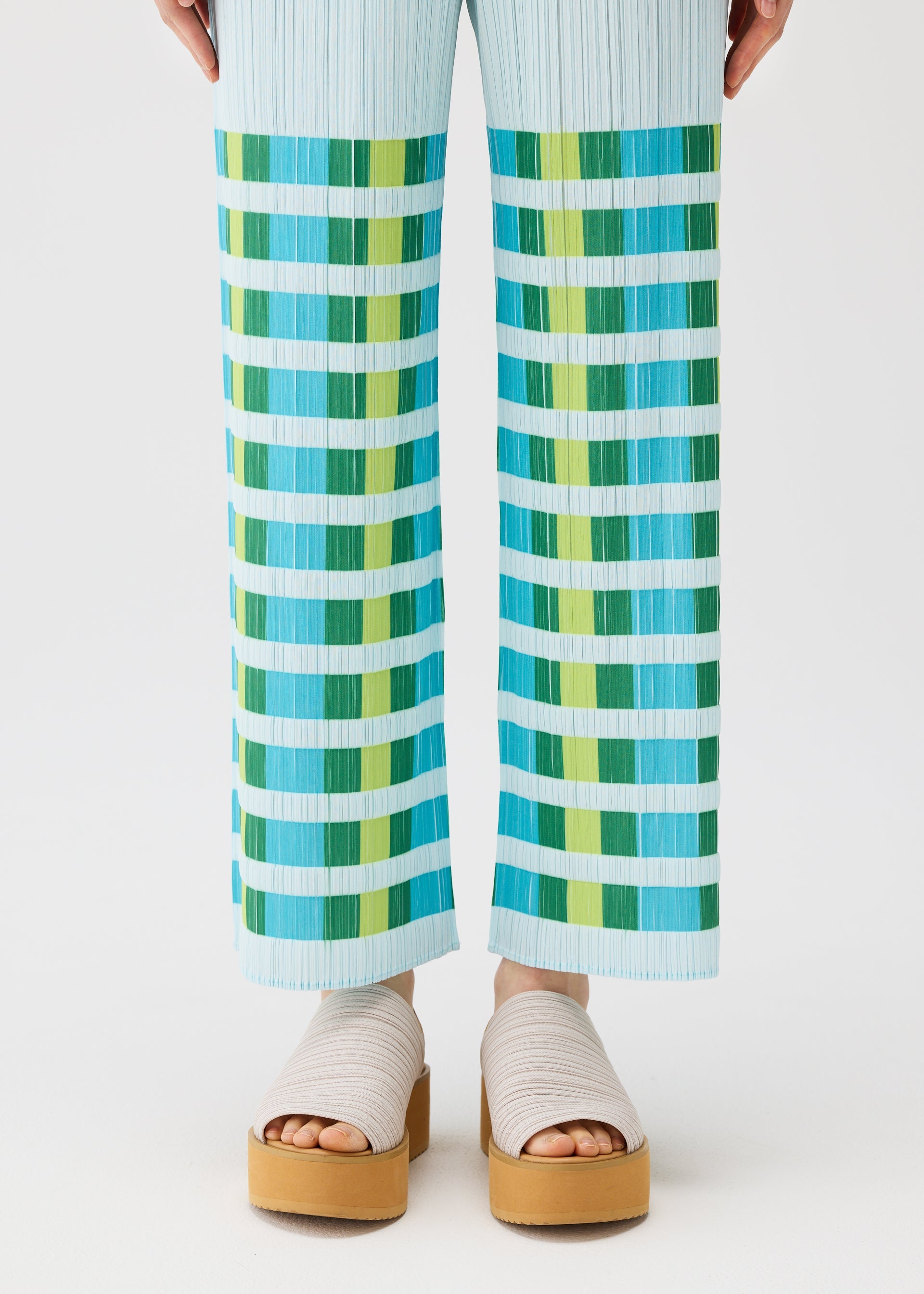Zoomed view A person wears the PLEATS PLEASE ISSEY MIYAKE BUMPY BASKET plaid straight pants with green and turquoise stripes, styled with beige platform sandals with thick straps on a white background.