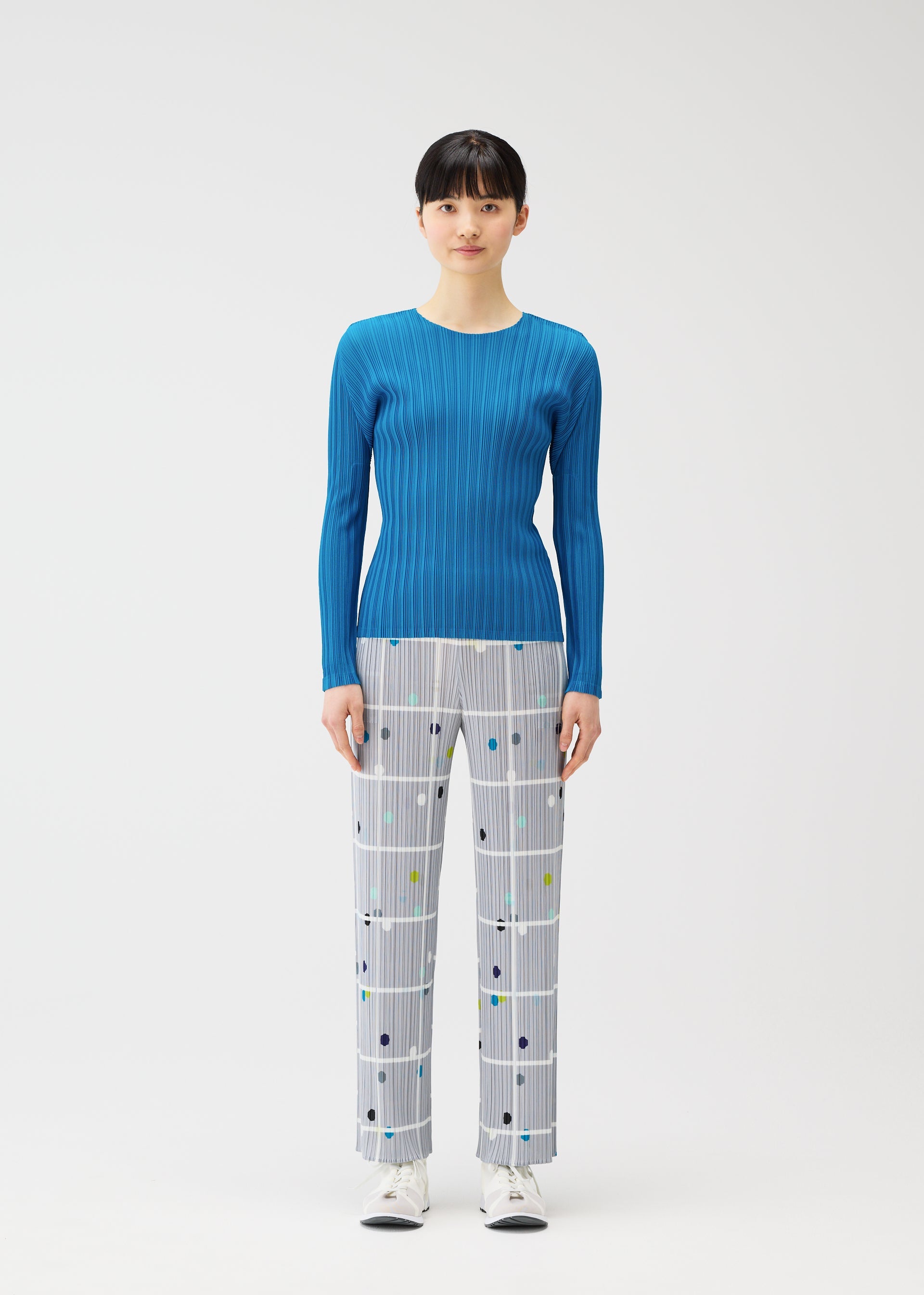 Zoomed view A woman faces forward in PLEATS PLEASE ISSEY MIYAKE's CROSSING DOTS straight pants with a plaid and dots grid pattern, a fitted blue ribbed long-sleeve top, and white sneakers against a plain white background.
