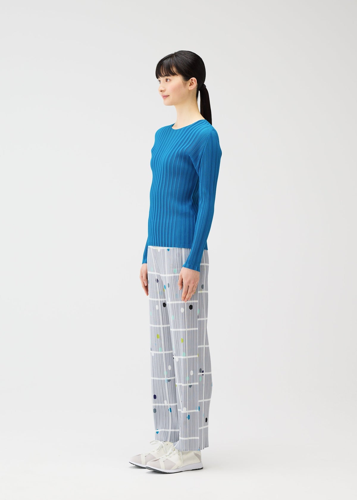 A woman in profile poses against a white background, dressed in the CROSSING DOTS pants from PLEATS PLEASE ISSEY MIYAKE, paired with a blue ribbed long-sleeve top and white shoes adorned with bows. view 4 of 5