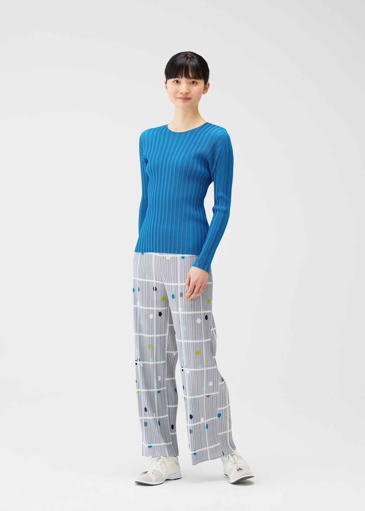 A woman stands against a plain white background wearing the CROSSING DOTS wide-leg straight pants by PLEATS PLEASE ISSEY MIYAKE, featuring a plaid and dots pattern, paired with a bright blue ribbed top and white sneakers. view 5 of 5