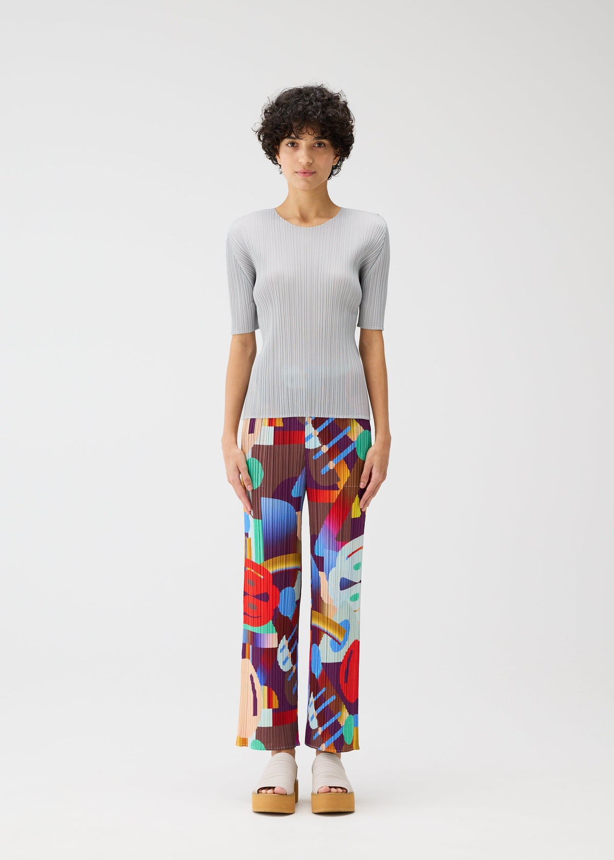 A person stands against a plain white background, wearing the playful PLAYGROUND straight pants with an abstract pattern and a light gray ribbed short-sleeve top by PLEATS PLEASE ISSEY MIYAKE, paired with platform sandals. view 4 of 7