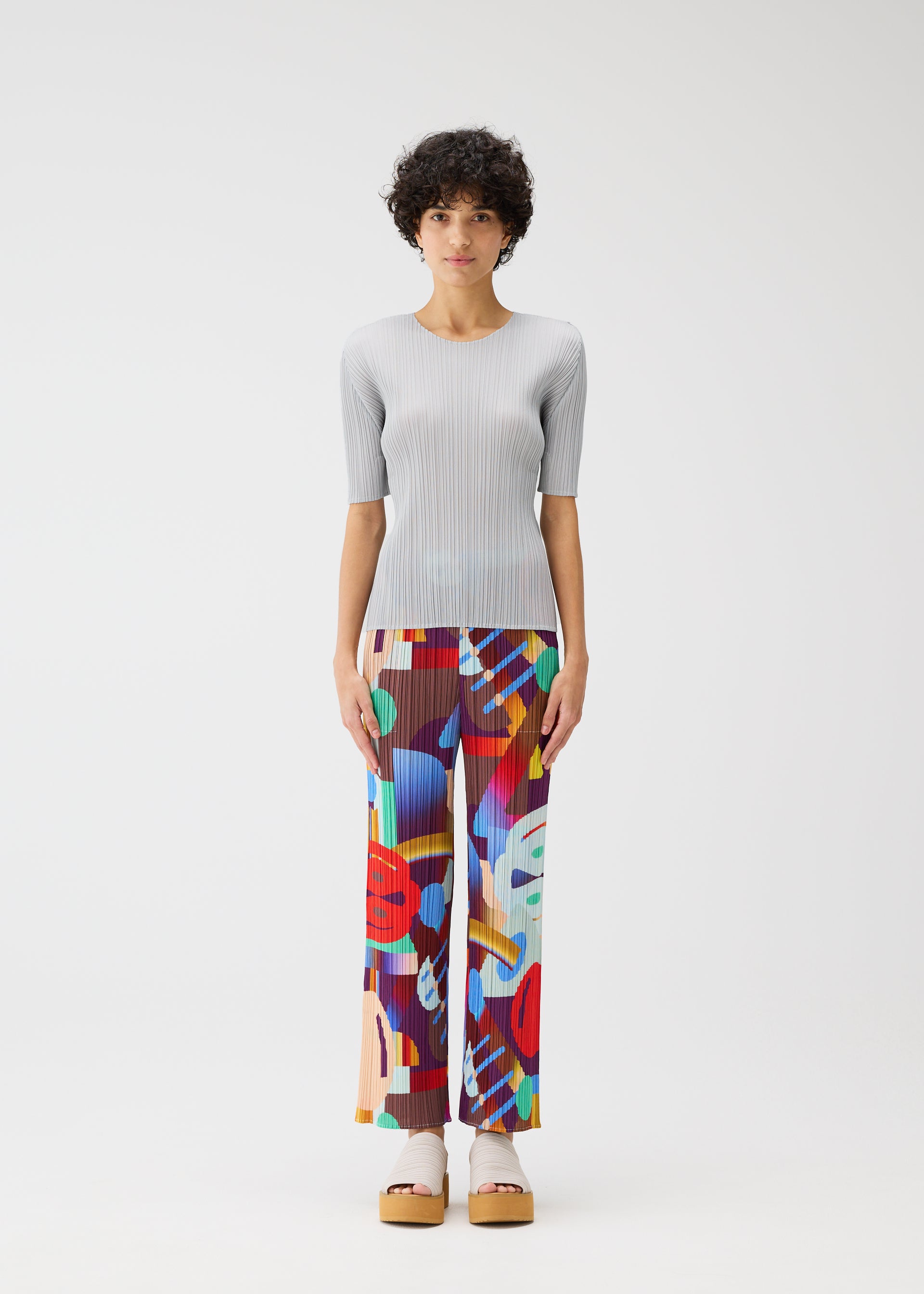 Zoomed view A person stands against a plain white background, wearing the playful PLAYGROUND straight pants with an abstract pattern and a light gray ribbed short-sleeve top by PLEATS PLEASE ISSEY MIYAKE, paired with platform sandals.
