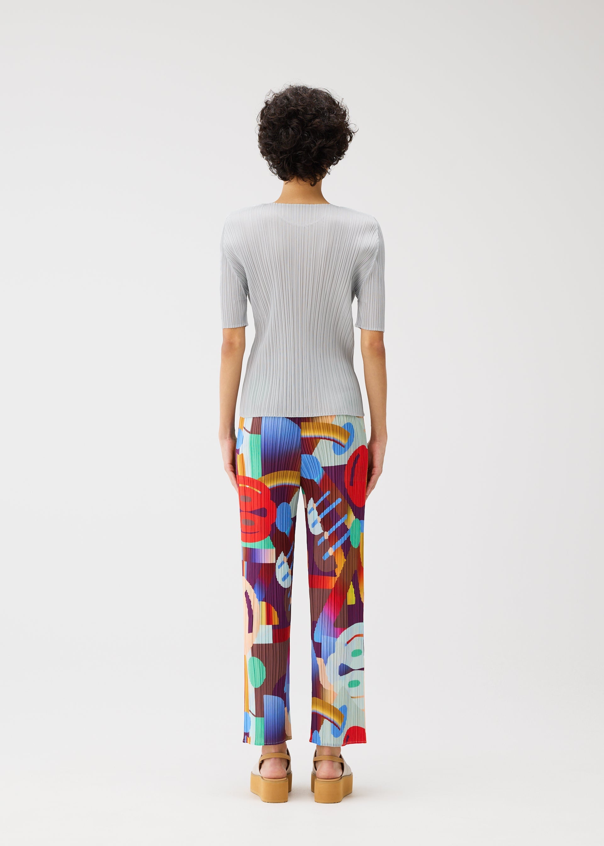 Zoomed view Facing away, a person with short curly hair wears the PLAYGROUND ribbed top and colorful geometric pants by PLEATS PLEASE ISSEY MIYAKE, styled with tan platform sandals against a plain white background.