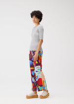 A person with short curly hair stands sideways against a white background, wearing the PLAYGROUND top and colorful geometric pants by PLEATS PLEASE ISSEY MIYAKE, paired with beige platform sandals.