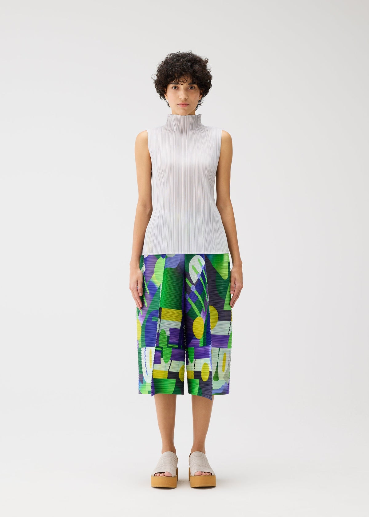 A person with short curly hair wears the PLAYGROUND sleeveless high-neck white pleated top and geometric culotte shorts by PLEATS PLEASE ISSEY MIYAKE, standing against a plain white background. view 5 of 8