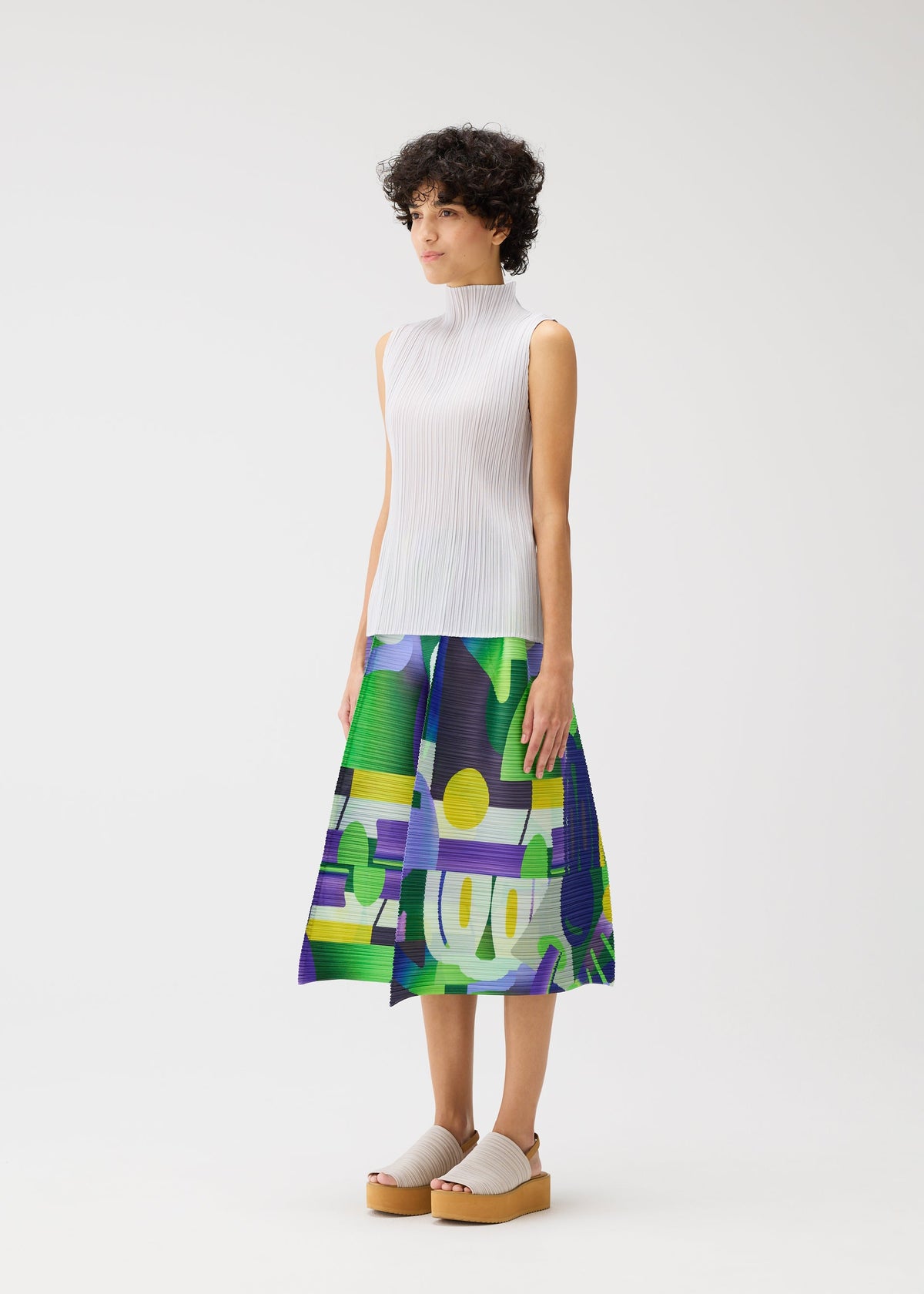 A woman with short curly hair models the PLAYGROUND midi skirt by PLEATS PLEASE ISSEY MIYAKE, featuring a playful green, blue, purple, and yellow print. She pairs it with a white ribbed sleeveless top and beige platform shoes against a white background. view 6 of 8