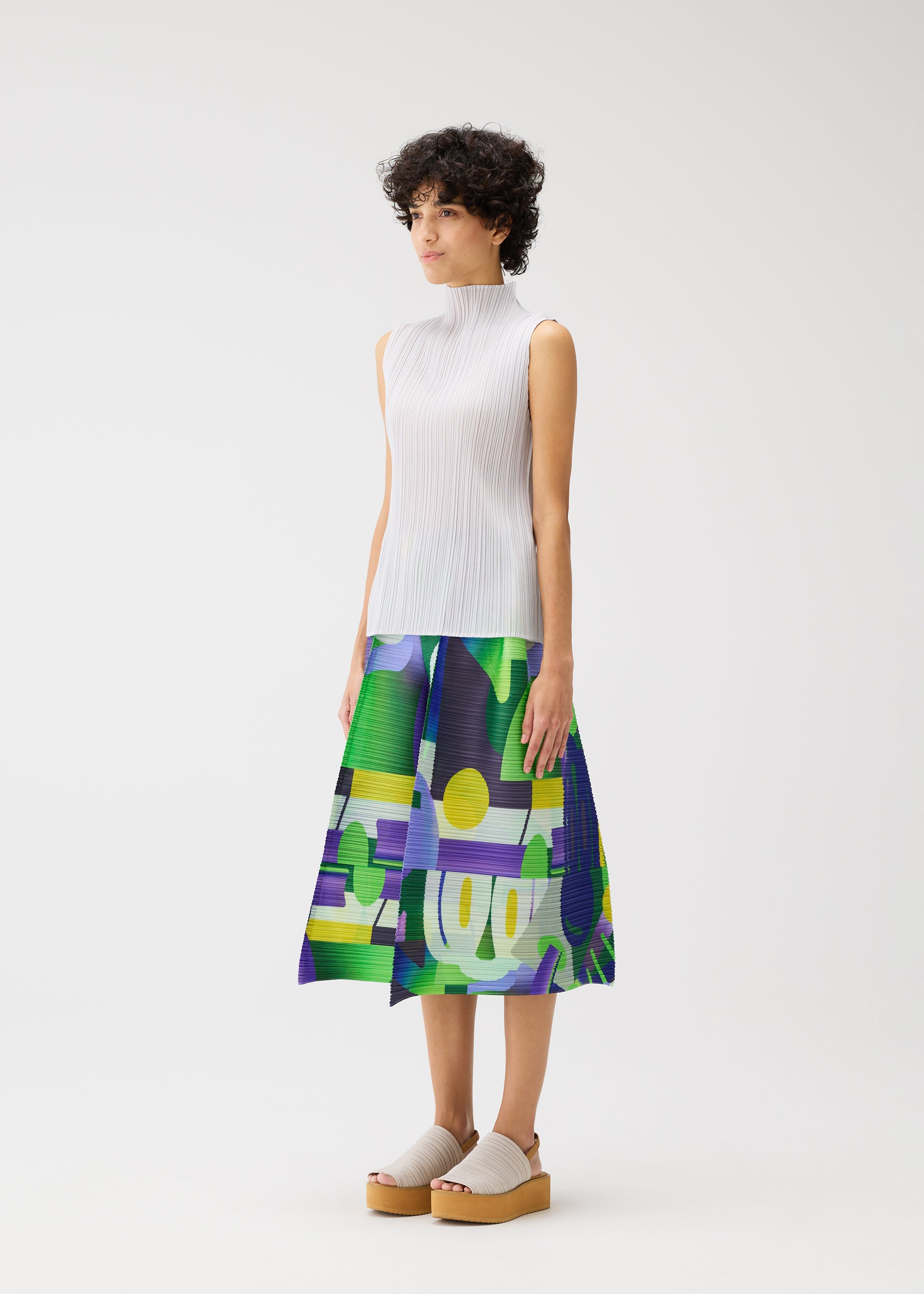 Zoomed view A woman with short curly hair models the PLAYGROUND midi skirt by PLEATS PLEASE ISSEY MIYAKE, featuring a playful green, blue, purple, and yellow print. She pairs it with a white ribbed sleeveless top and beige platform shoes against a white background.