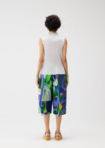 A person with short curly hair faces away, wearing the PLAYGROUND sleeveless white pleated top and wide geometric-print pants in green, purple, and blue by PLEATS PLEASE ISSEY MIYAKE, paired with light brown platform sandals against a white background.