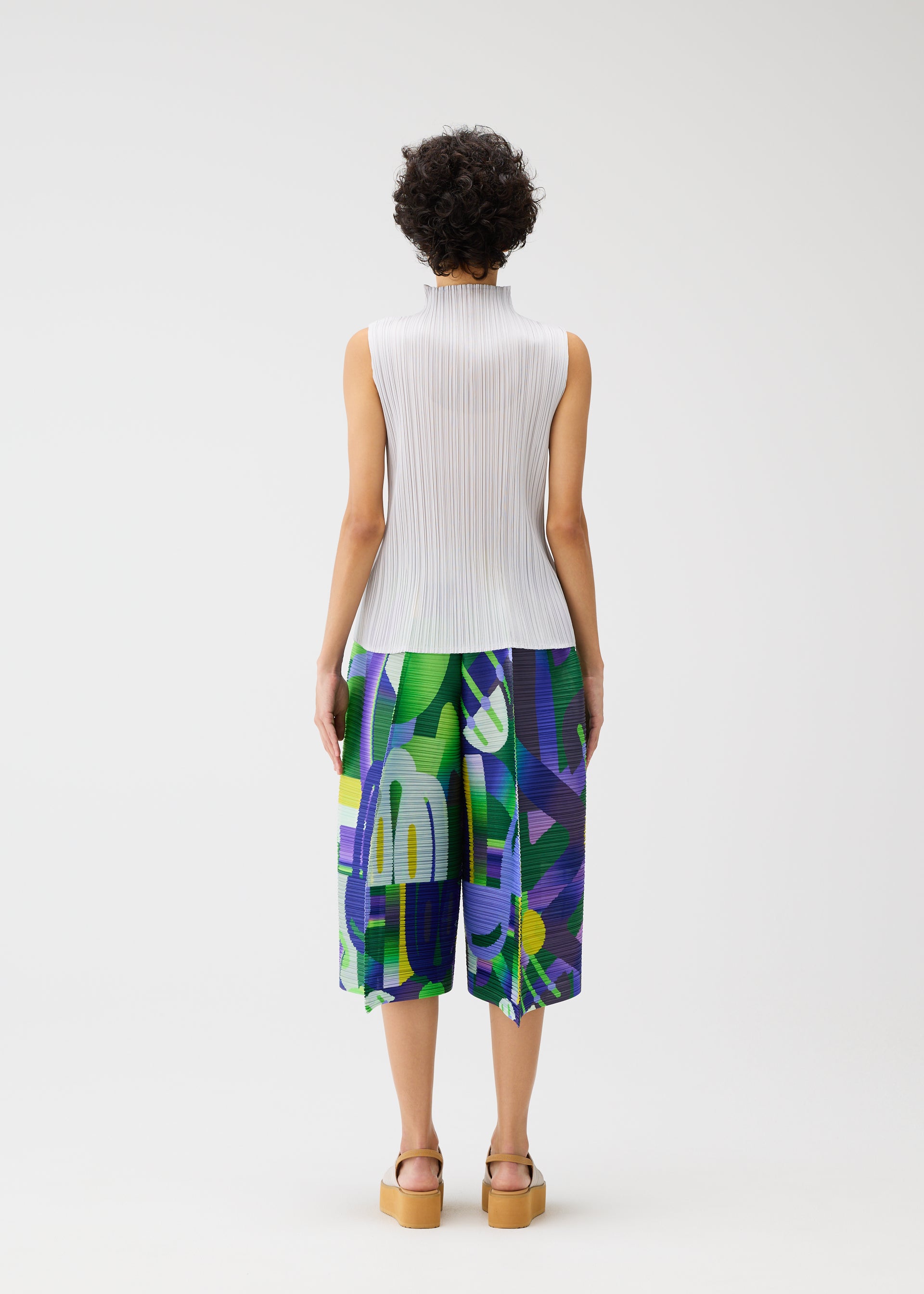 Zoomed view A person with short curly hair faces away, wearing the PLAYGROUND sleeveless white pleated top and wide geometric-print pants in green, purple, and blue by PLEATS PLEASE ISSEY MIYAKE, paired with light brown platform sandals against a white background.