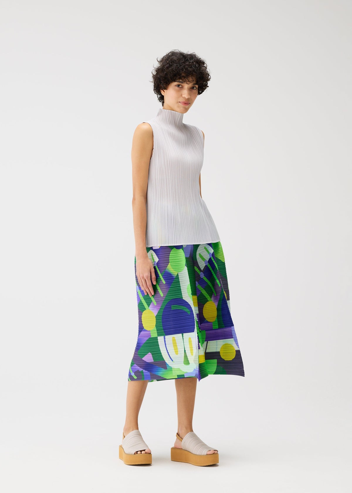 A woman with short curly hair models the PLAYGROUND pleated top and playful printed midi skirt from PLEATS PLEASE ISSEY MIYAKE, styled with neutral platform sandals against a plain white background. view 8 of 8