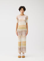 A person with short curly hair stands facing forward in the PLEATS PLEASE ISSEY MIYAKE SEA BUBBLE set—matching ribbed knit top and straight pants in soft beachy blue-beige tones—paired with light platform sandals, against a plain white background.
