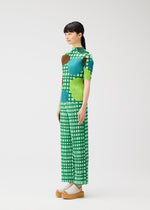 A woman stands sideways in PLEATS PLEASE ISSEY MIYAKE's BASKET outfit featuring a green and white block check print with blue and brown circles, short sleeves, straight pants, and beige platform sandals against a plain white background.