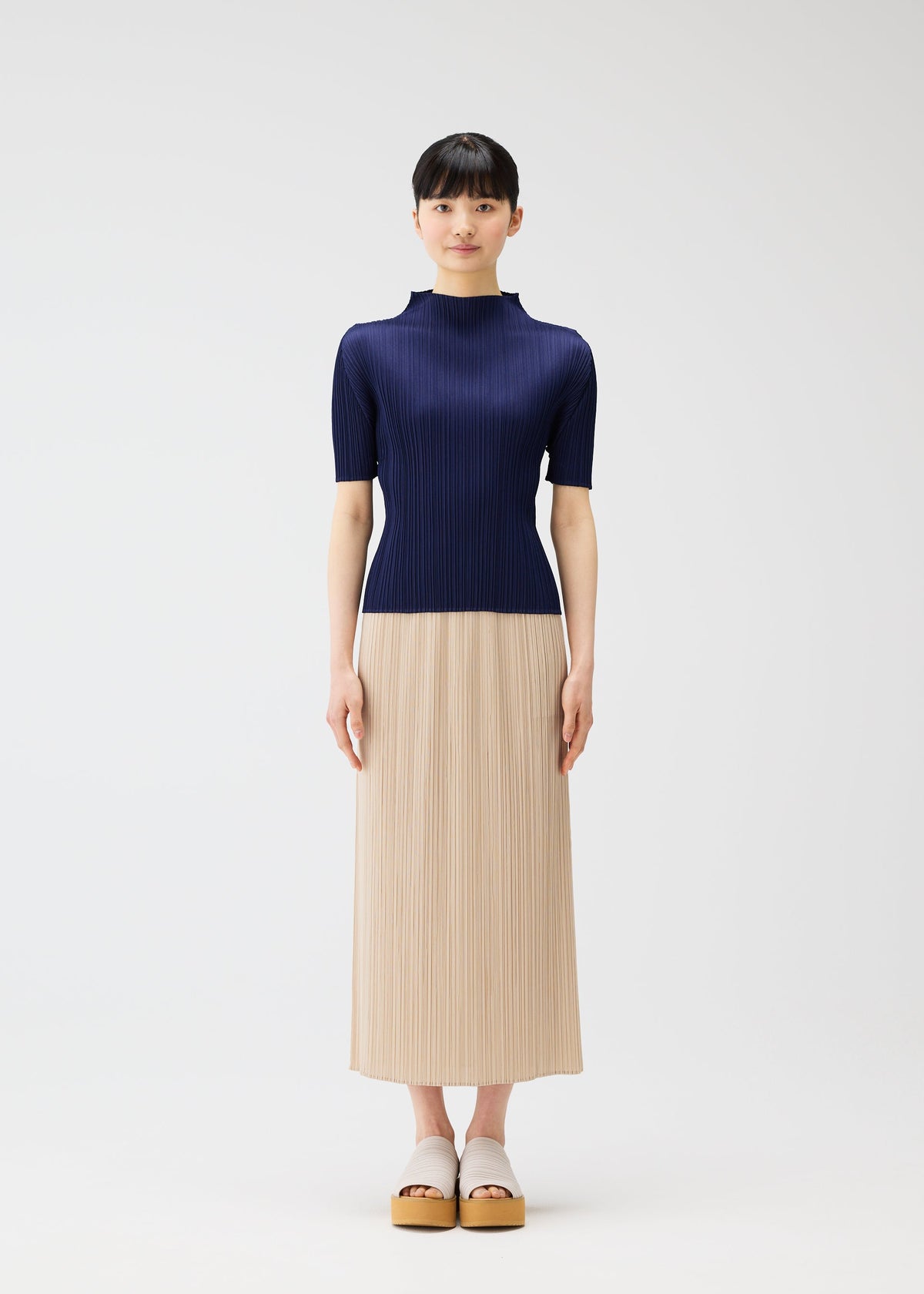 A woman faces forward against a plain light background, wearing a navy ribbed top and beige pleated skirt from the MONTHLY COLORS : MAY collection by PLEATS PLEASE ISSEY MIYAKE, paired with beige platform sandals. view 8 of 11