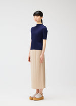 A woman in a minimalist studio wears the MONTHLY COLORS : MAY collection by PLEATS PLEASE ISSEY MIYAKE: a short-sleeve navy top, beige beach-inspired skirt, and light platform sandals. She smiles softly, looking to the side with straight dark hair and bangs.