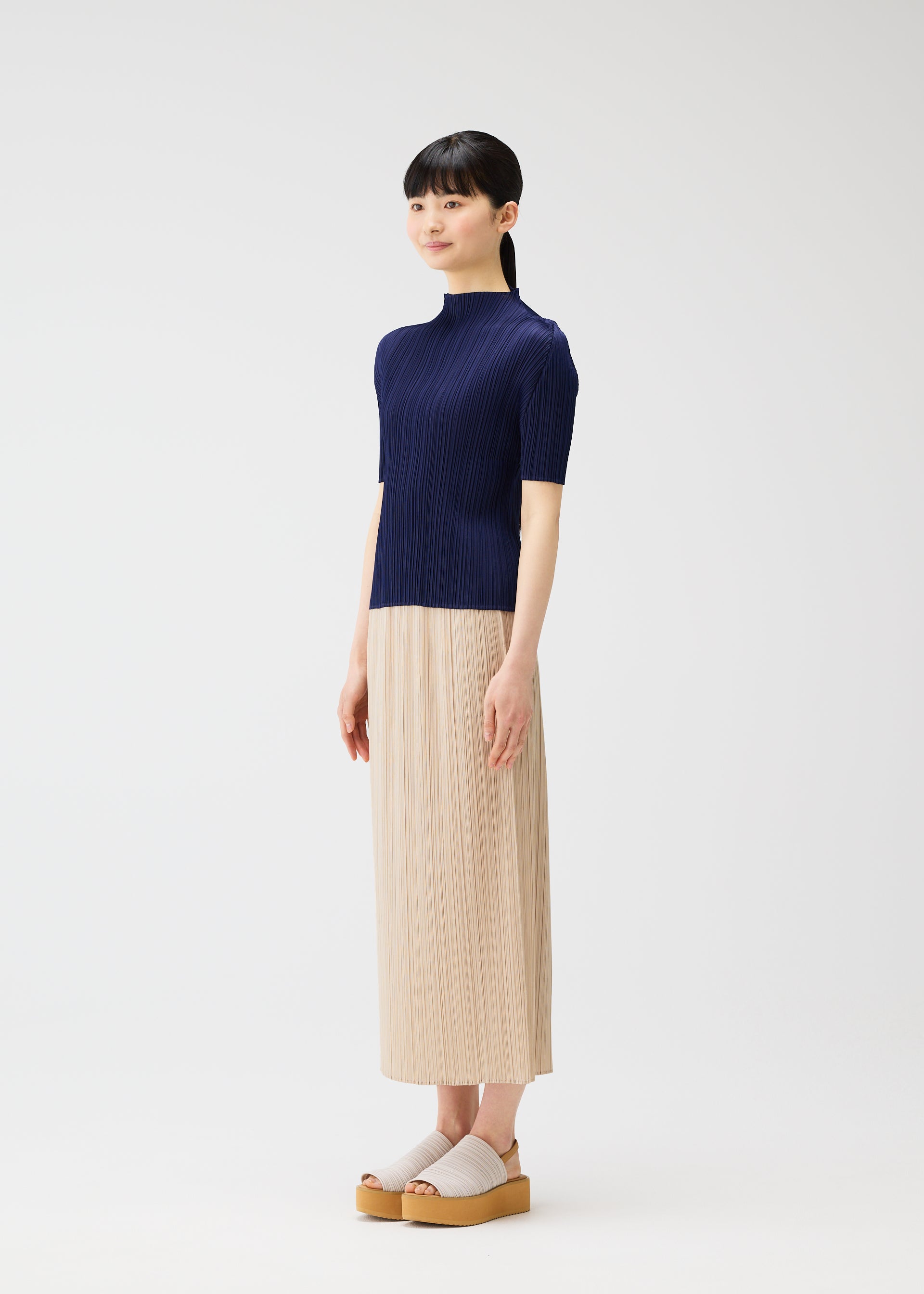 Zoomed view A woman in a minimalist studio wears the MONTHLY COLORS : MAY collection by PLEATS PLEASE ISSEY MIYAKE: a short-sleeve navy top, beige beach-inspired skirt, and light platform sandals. She smiles softly, looking to the side with straight dark hair and bangs.
