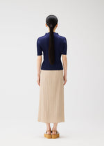 A woman with long black hair in a ponytail stands facing away, wearing the MONTHLY COLORS : MAY navy top and beige summer skirt by PLEATS PLEASE ISSEY MIYAKE, with tan platform sandals, against a white background.