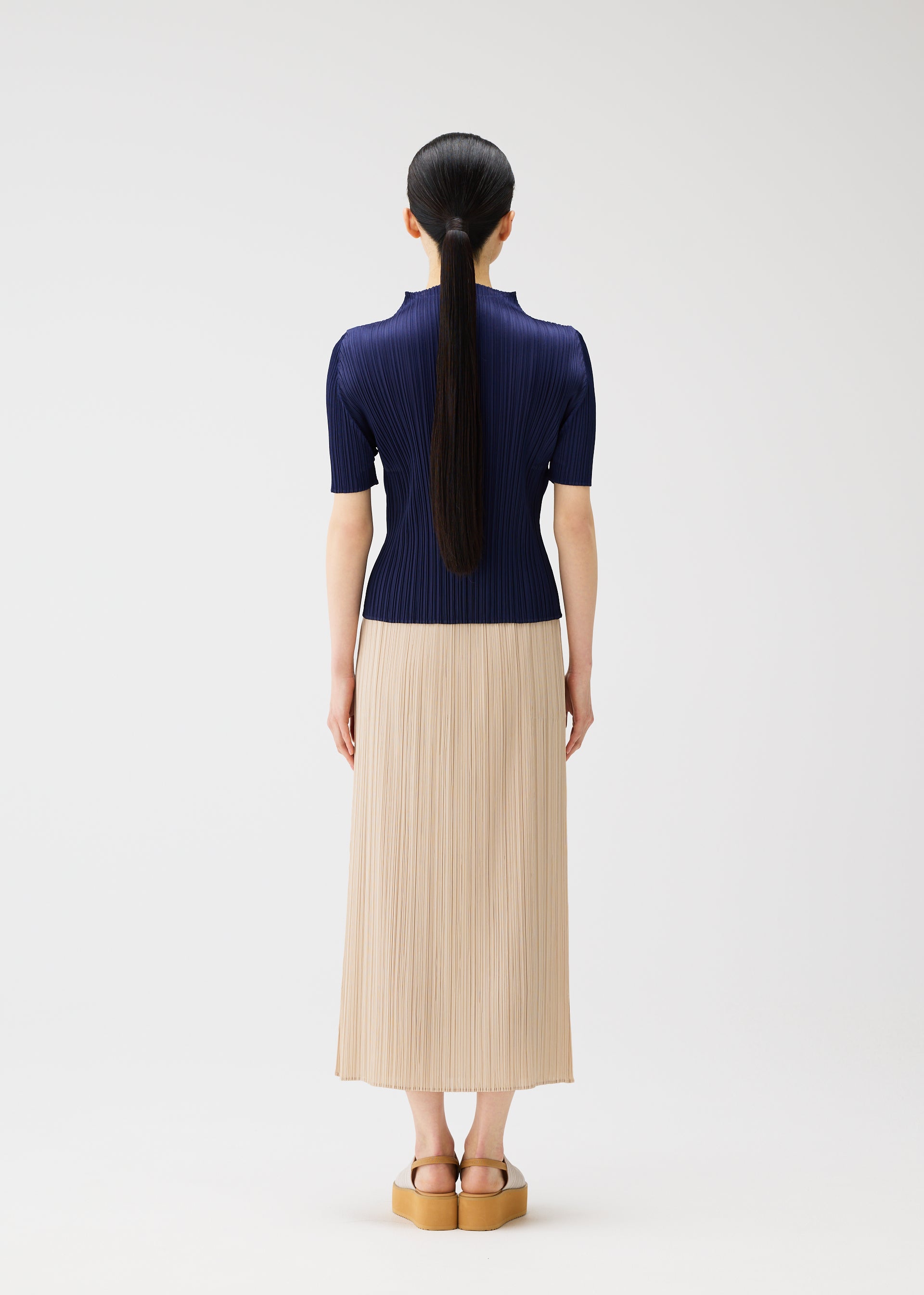 Zoomed view A woman with long black hair in a ponytail stands facing away, wearing the MONTHLY COLORS : MAY navy top and beige summer skirt by PLEATS PLEASE ISSEY MIYAKE, with tan platform sandals, against a white background.