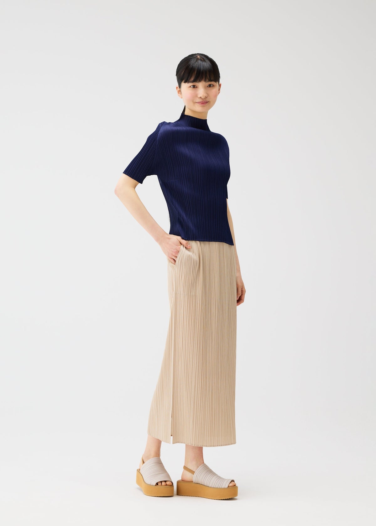 A woman with straight dark hair and bangs smiles softly, wearing the "MONTHLY COLORS : MAY" collection by PLEATS PLEASE ISSEY MIYAKE—featuring a navy short-sleeve top, a pleated beach-inspired skirt, and light beige platform sandals. view 11 of 11