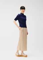 A woman with straight dark hair and bangs smiles softly, wearing the "MONTHLY COLORS : MAY" collection by PLEATS PLEASE ISSEY MIYAKE—featuring a navy short-sleeve top, a pleated beach-inspired skirt, and light beige platform sandals.