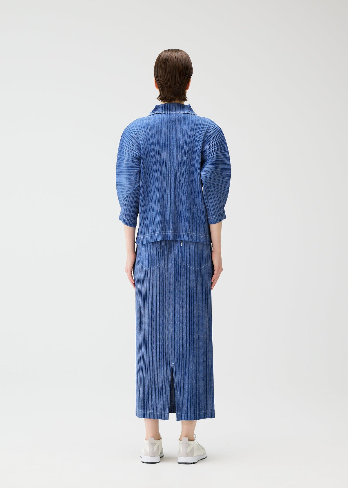A person with short brown hair wears PLEATS PLEASE ISSEY MIYAKE’s COTTON DENIM in ocean breeze—a ribbed, calf-length skirt and matching jacket with elbow-length sleeves—paired with white sneakers against a plain background. view 7 of 8