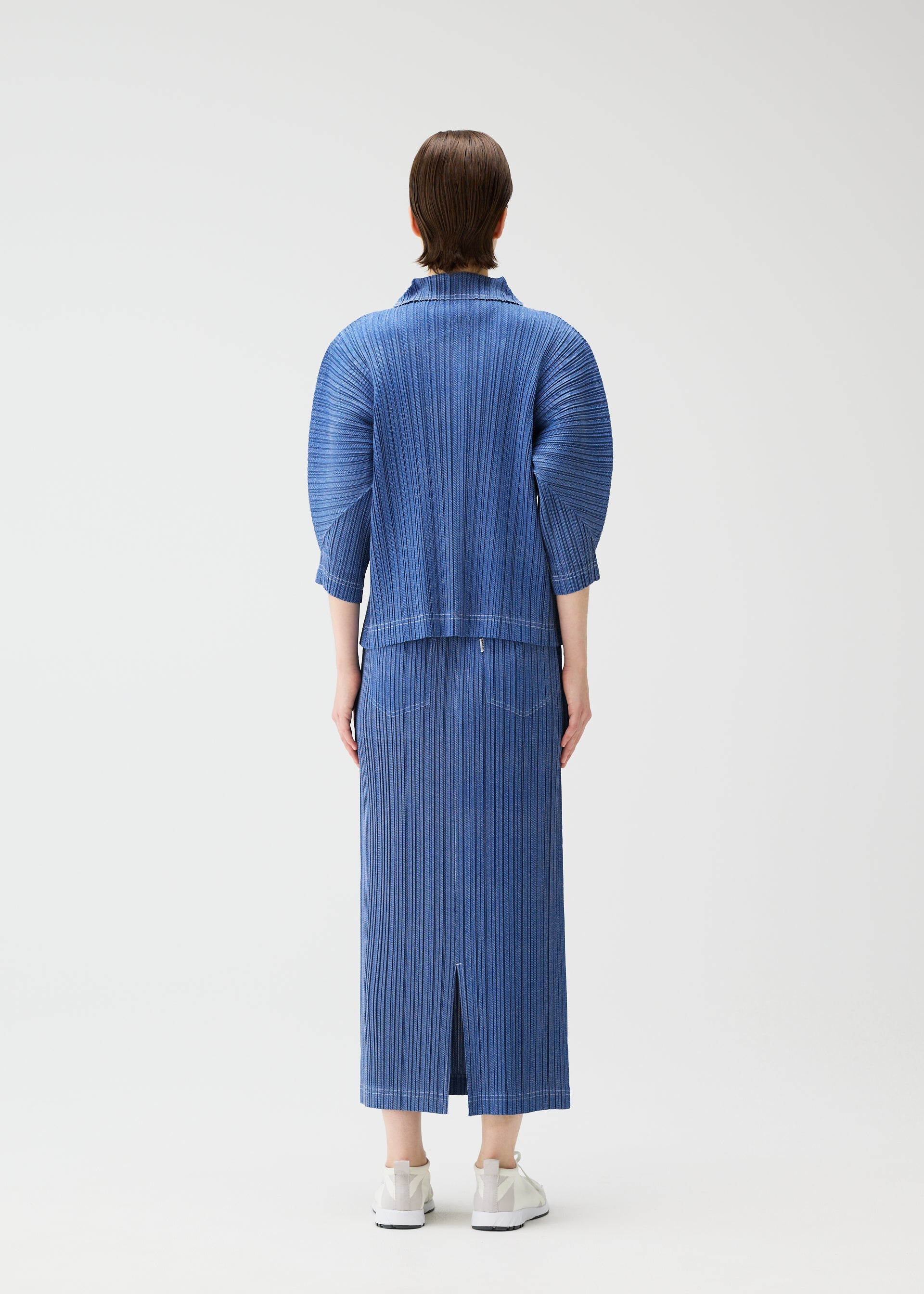 Zoomed view A person with short brown hair wears PLEATS PLEASE ISSEY MIYAKE’s COTTON DENIM in ocean breeze—a ribbed, calf-length skirt and matching jacket with elbow-length sleeves—paired with white sneakers against a plain background.