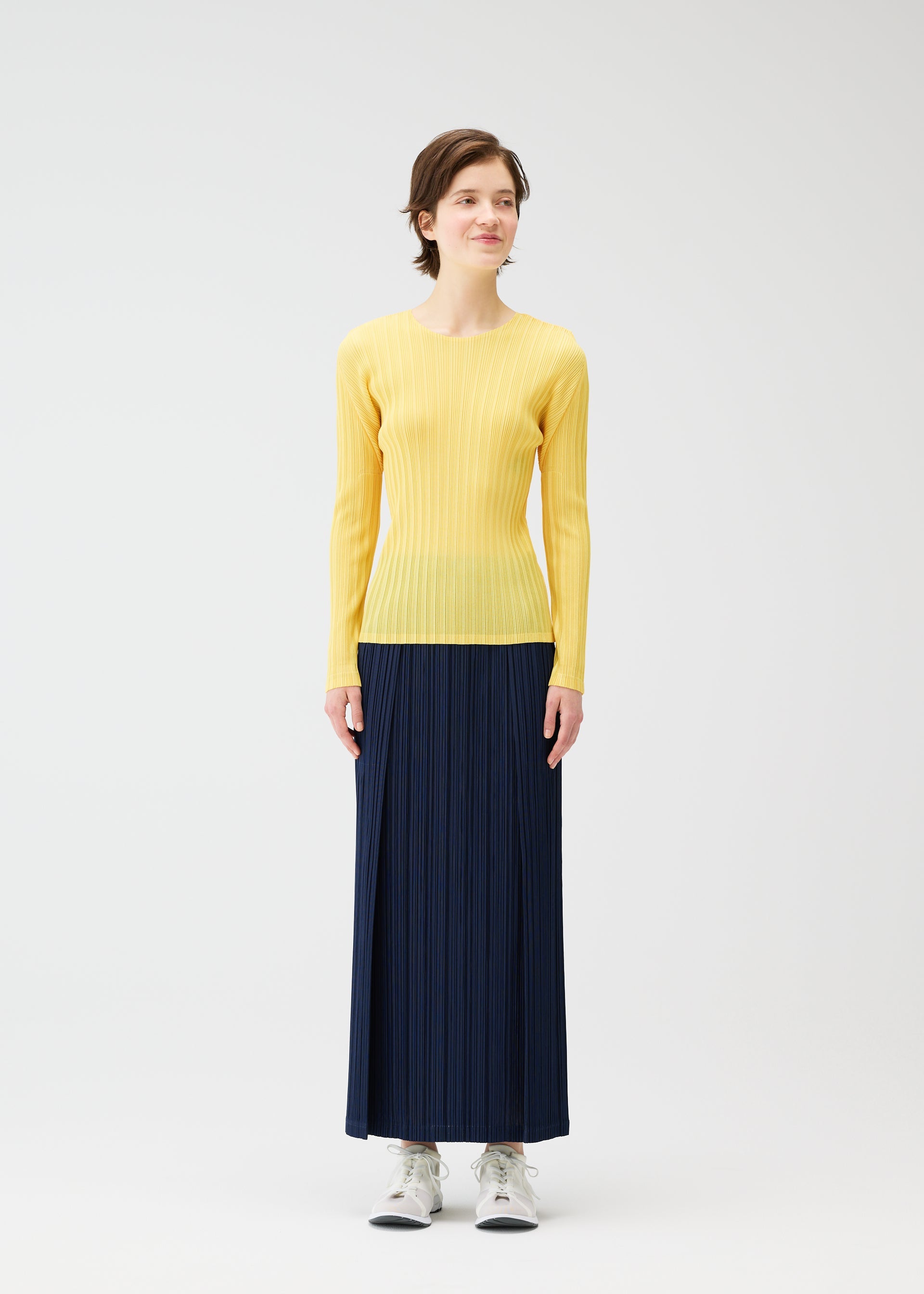 Zoomed view A person smiles gently, looking to the side, wearing a yellow ribbed long-sleeve top tucked into PLEATS PLEASE ISSEY MIYAKE THICKER BOTTOMS 1 navy pleated skirt with pockets, paired with white sneakers against a plain white background.