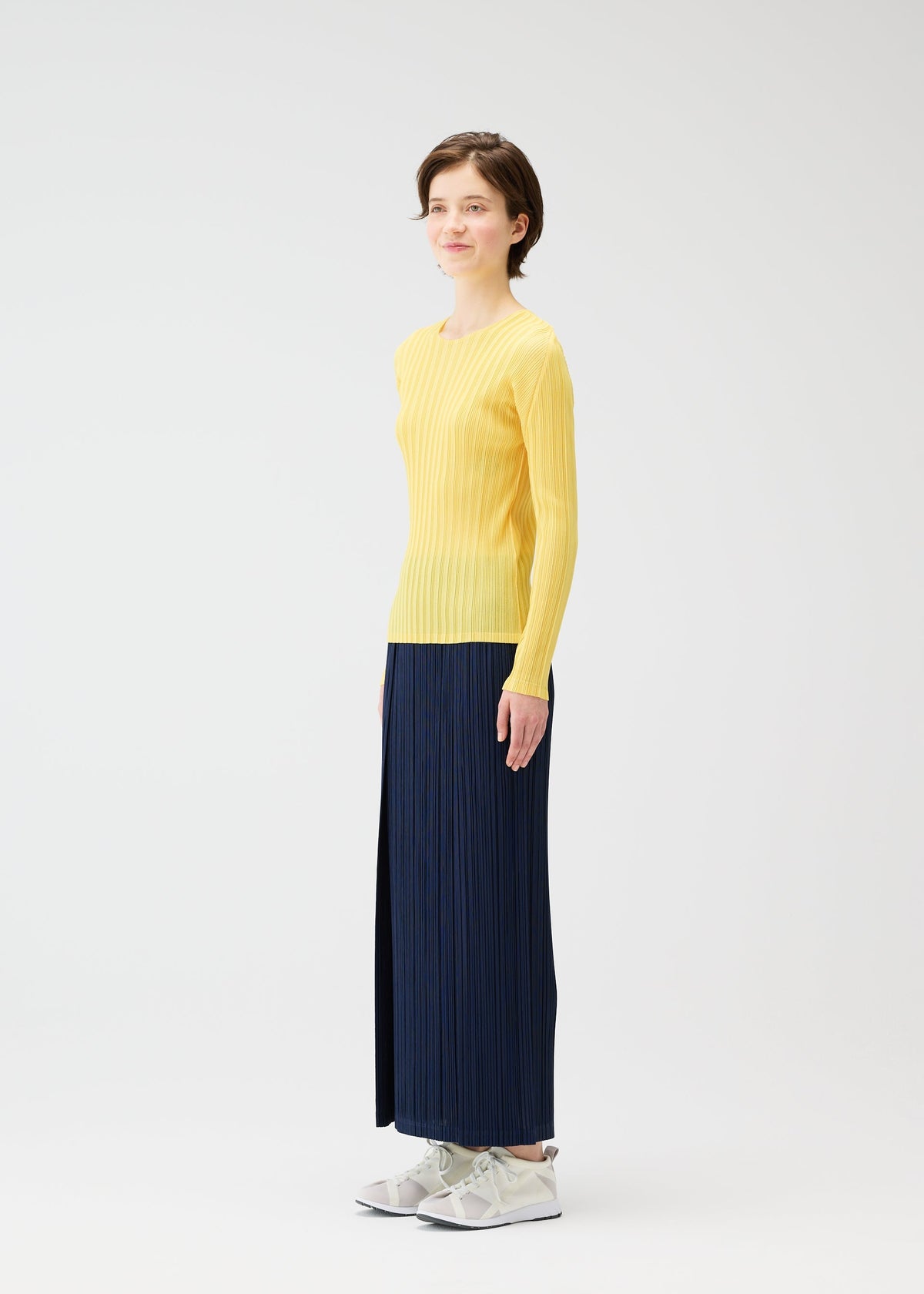 A person stands against a plain white background, wearing PLEATS PLEASE ISSEY MIYAKE's THICKER BOTTOMS 1 with a long-sleeved yellow ribbed top and white sneakers, looking forward with a slight smile. view 9 of 11