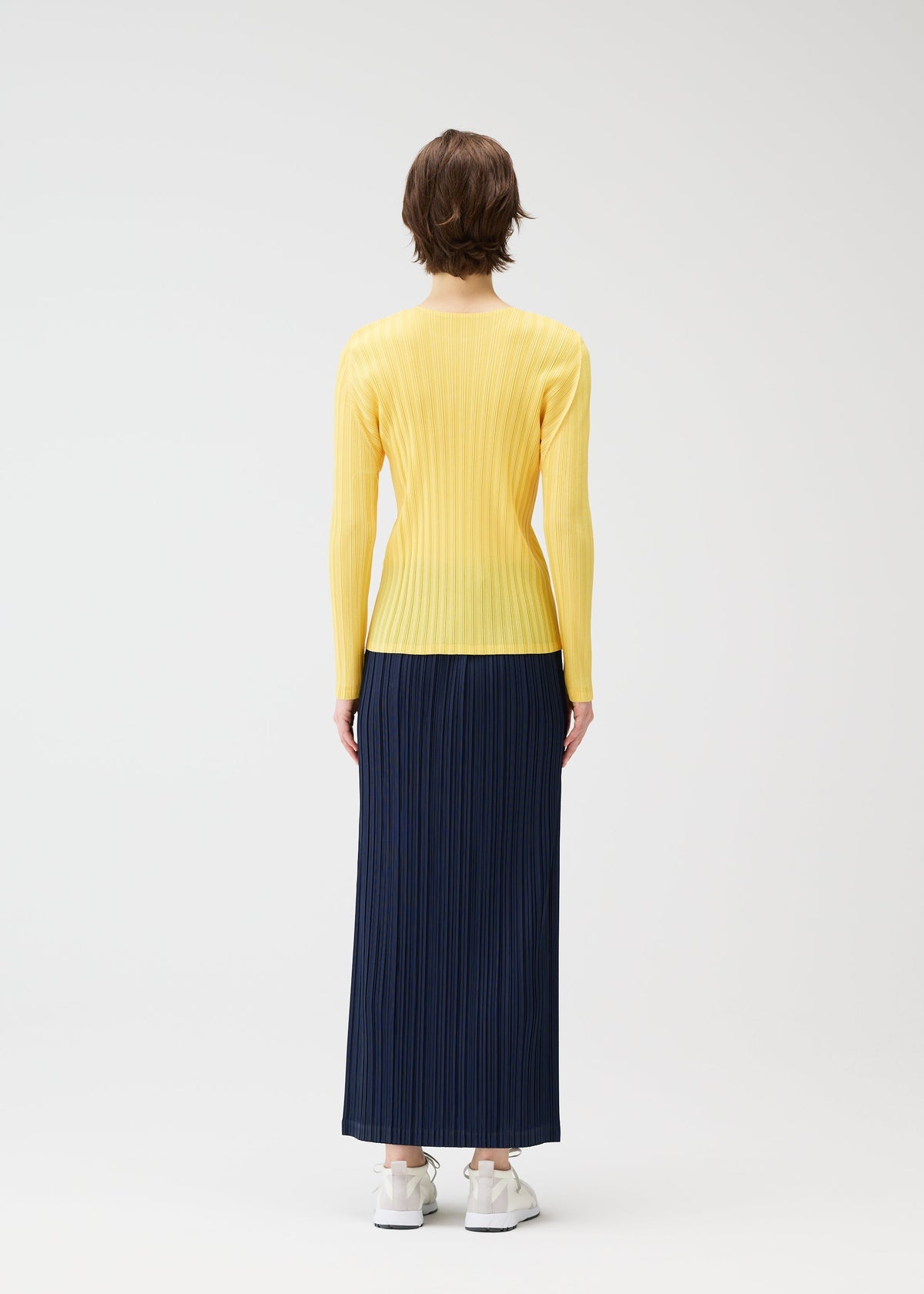 A person with short brown hair faces away, wearing a long-sleeve yellow ribbed top tucked into PLEATS PLEASE ISSEY MIYAKE's THICKER BOTTOMS 1, a navy blue pleated maxi skirt with pockets, and white sneakers against a plain white background. view 10 of 11