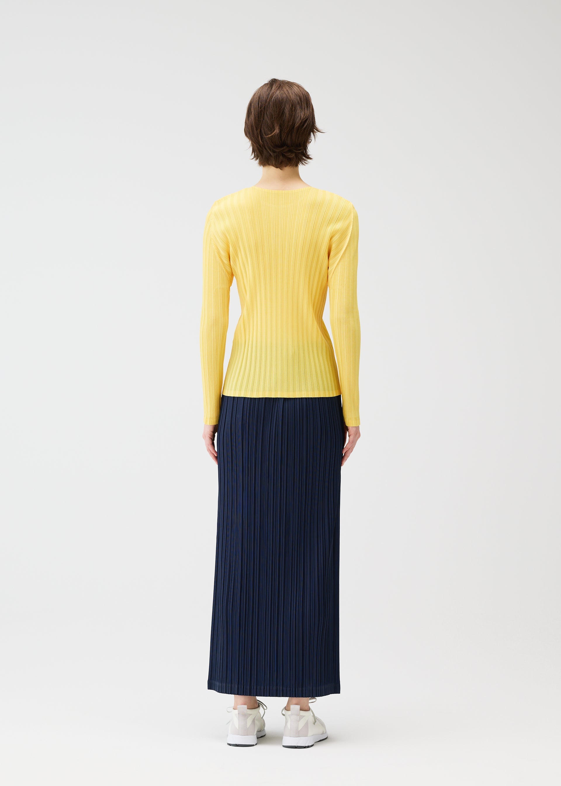 Zoomed view A person with short brown hair faces away, wearing a long-sleeve yellow ribbed top tucked into PLEATS PLEASE ISSEY MIYAKE's THICKER BOTTOMS 1, a navy blue pleated maxi skirt with pockets, and white sneakers against a plain white background.