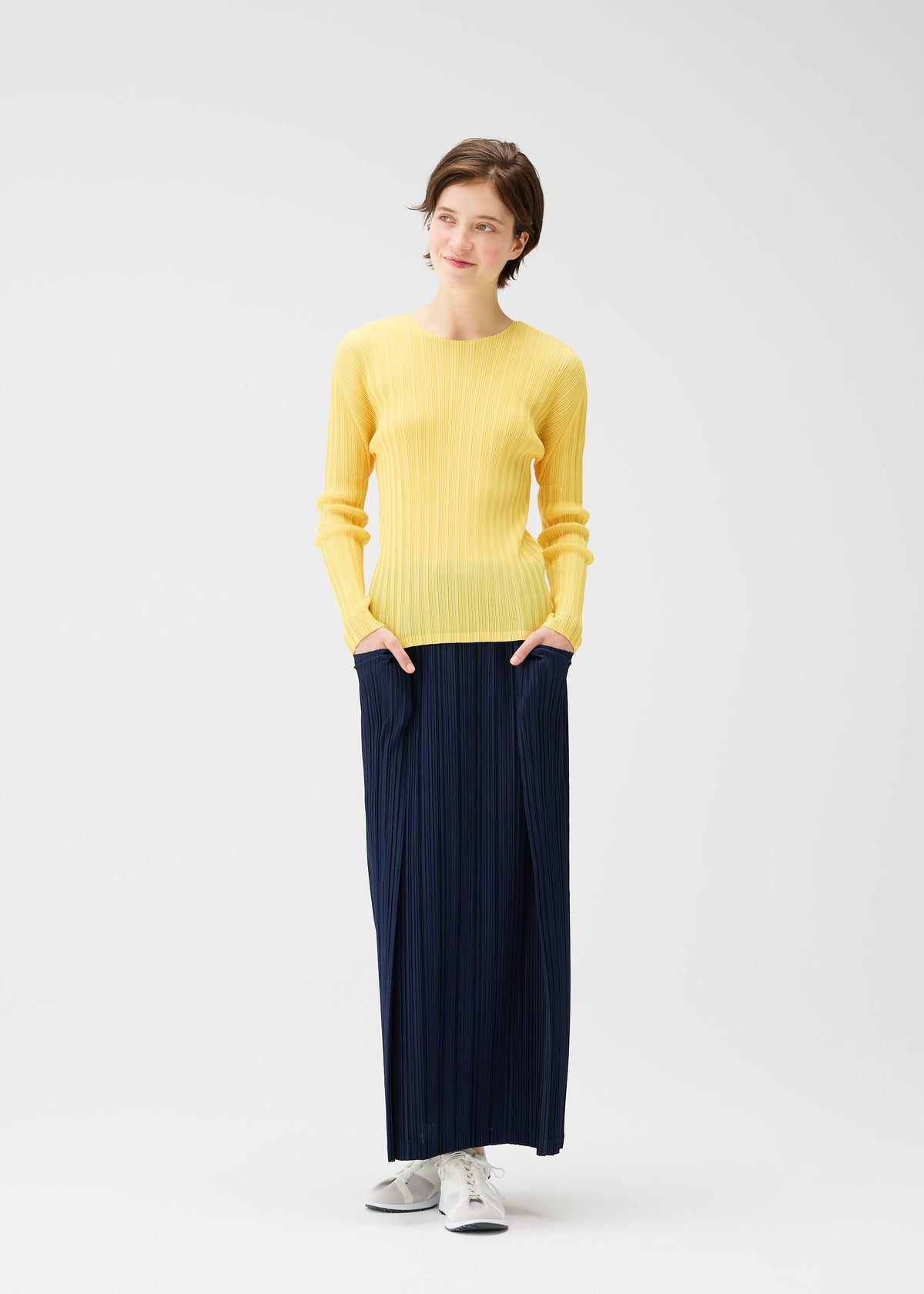 A woman stands against a white background, wearing a yellow sweater, white sneakers, and the PLEATS PLEASE ISSEY MIYAKE THICKER BOTTOMS 1 navy skirt with pockets, her hands in her pockets and a slight smile on her face. view 11 of 11