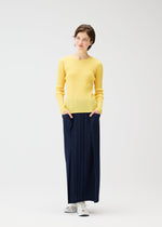 A woman stands against a white background, wearing a yellow sweater, white sneakers, and the PLEATS PLEASE ISSEY MIYAKE THICKER BOTTOMS 1 navy skirt with pockets, her hands in her pockets and a slight smile on her face.