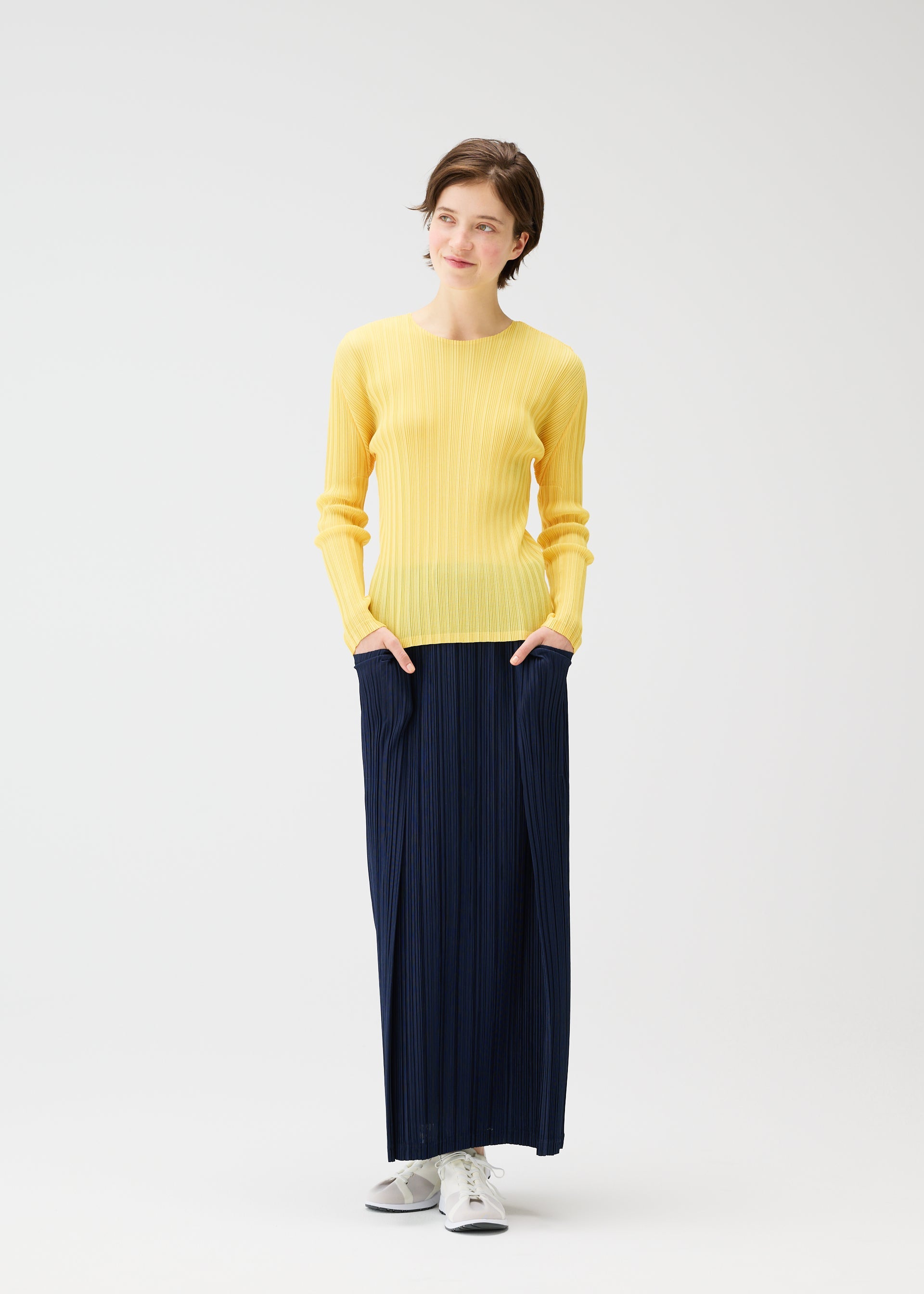 Zoomed view A woman stands against a white background, wearing a yellow sweater, white sneakers, and the PLEATS PLEASE ISSEY MIYAKE THICKER BOTTOMS 1 navy skirt with pockets, her hands in her pockets and a slight smile on her face.