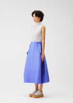 Against a white background, a person in PLEATS PLEASE ISSEY MIYAKE’s BOUNCY BASKET blue pleated midi skirt with pockets, a sleeveless white top, and light gray platform sandals showcases modern women's style with short, curly hair.