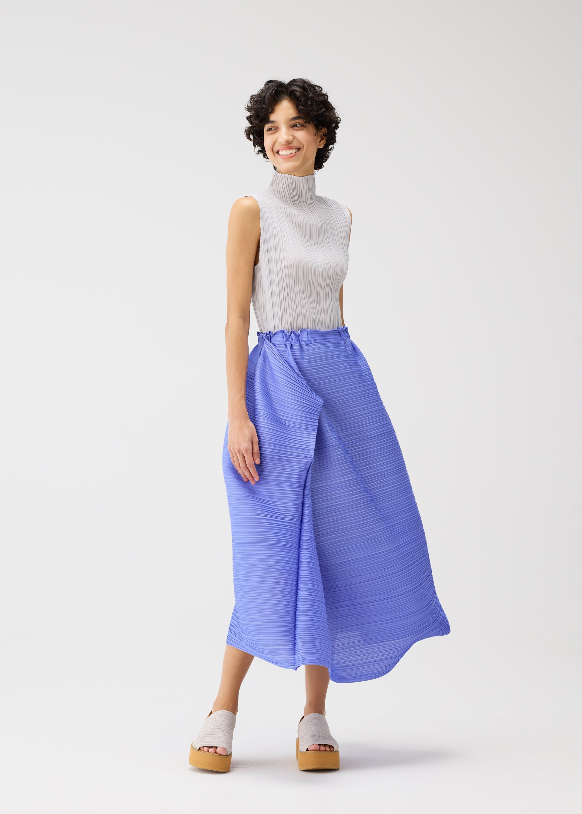 Zoomed view A woman with short curly hair models the BOUNCY BASKET by PLEATS PLEASE ISSEY MIYAKE, wearing a sleeveless high-neck white top and blue pleated skirt with platform sandals against a plain white background.