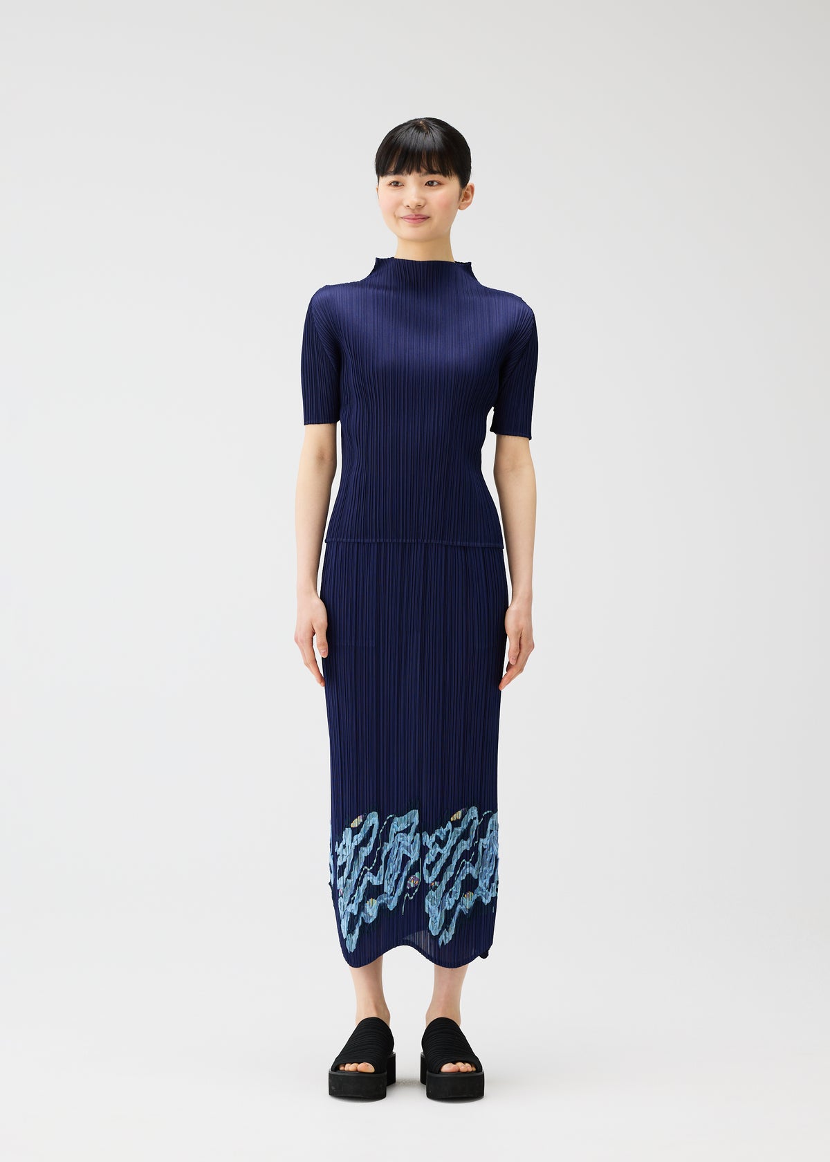 A woman faces forward, smiling softly, wearing the PLEATS PLEASE ISSEY MIYAKE I SPY! navy dress with pleated, embroidered skirt and light blue patterns at the hem. She pairs it with black platform sandals against a plain white background. view 5 of 10
