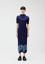 A woman faces forward, smiling softly, wearing the PLEATS PLEASE ISSEY MIYAKE I SPY! navy dress with pleated, embroidered skirt and light blue patterns at the hem. She pairs it with black platform sandals against a plain white background.